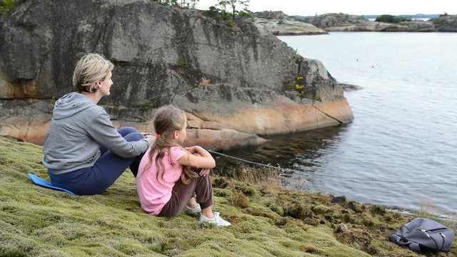 Mother and daughter sitting on the big stones on the sea shore, looking at water