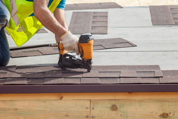 Construction worker putting the asphalt roofing (shingles) with nail gun on a new frame house