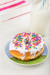 A frosted donut/doughnut with rainbow sprinkles . Milk and colorful napkins in the background