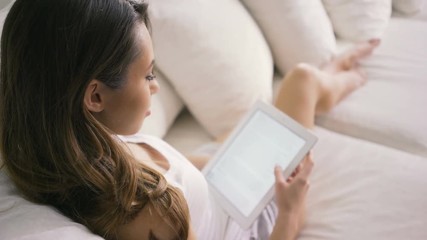 Young woman sitting on couch at home and reading ebook on tablet computer - Powered by Adobe