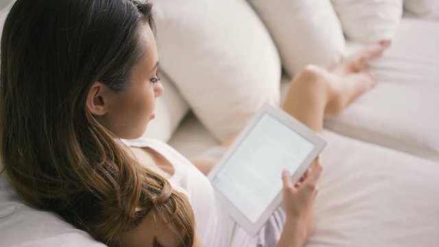 Young woman sitting on couch at home and reading ebook on tablet computer