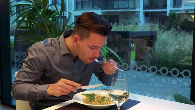 A young, handsome man sits at a restaurant and eats pasta, a glass of wine in front of him
