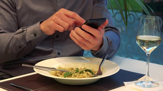 A man sits at a table in a restaurant and works on a smartphone, his meal in front of him