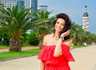 Fototapeta premium Woman corrects hair and smile in the long red dress. Pretty girl surrounded by tropical palms near sea