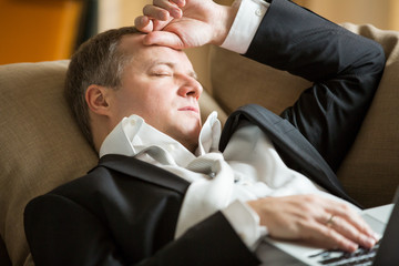 Businessman in suit lying on a couch with two cellphones and laptops, sleeping. Exhausted man relaxing in office early morning. Responsible executive working fell asleep. 