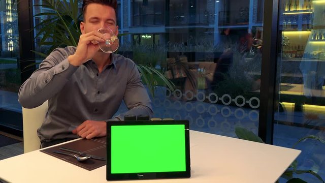A young, handsome man sits at a table in a restaurant, drinks wine and smiles at the camera - a tablet with a green screen in the foreground on the table
