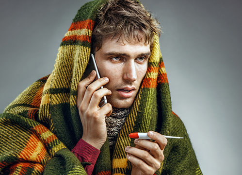 Sick man wrapped in blanket with a high temperature calling on the phone. Man suffering cold and winter flu virus. Health care concept