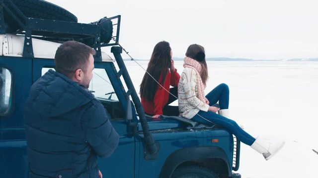 Three friends having a conversation on a hood of an old 4x4 off-road vehicle near the frozen lake. Winter nature in the background. 4K UHD 60 FPS RAW edited footage