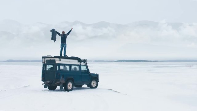 WIDE Caucasian male standing on the roof of old 4x4 off-road vehicle, spreading hands and enjoying the view of mountains over large lake. 4K UHD 60 FPS RAW edited footage