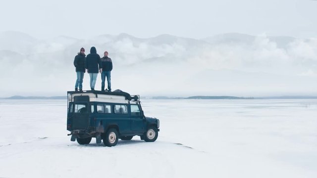 Three male friends standing on the roof of old 4x4 off-road vehicle during winter road trip, enjoying the view of mountains over large lake. 4K UHD 60 FPS RAW edited footage