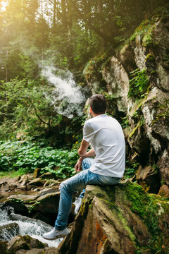 Smoker sitting on a rock Carpathian River, smoking an electronic cigarette, enjoying the beautiful nature