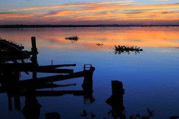 Beautiful sunset on the river parana in entre rios, Argentina, south america. An old wooden pier, the sky in orange tones is reflected on the water and aquatic plants pass by the river. - obrazy, fototapety, plakaty