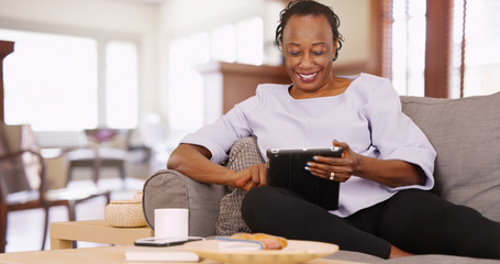 An elderly black woman uses her tablet while relaxing on the couch - Powered by Adobe