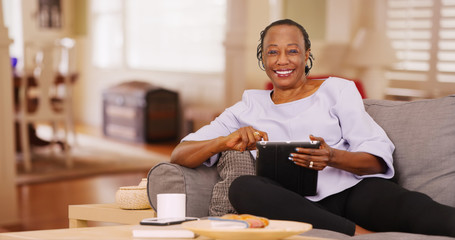 An elderly black woman happily uses her tablet while looking at the camera