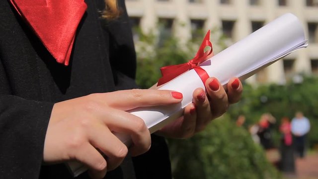 Close-up of female hands holding diploma with red ribbon, graduation ceremony