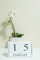 Closeup white wooden calendar with black 15 february word with white orchid flower on white wood desk and cream color wallpaper in room textured background in selective focus at the calendar