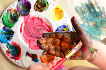 Child painting her hand with a paintbrush and background with palette of colorful paint
