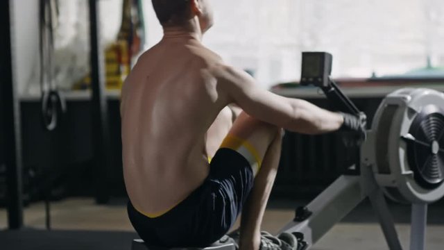 Tracking of sporty shirtless man working out on rowing machine and building up his upper body muscles 
