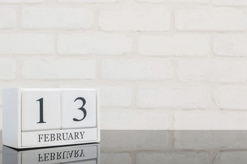 Closeup white wooden calendar with black 13 february word on black glass table and white brick wall textured background with copy space in selective focus at the calendar