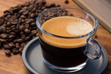 A transparent glass cup of hot espresso on a saucer standing on a wooden table top, surrounded by coffee beans and a vintage book