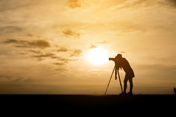 Silhouette of a photographer,she use a tripod,she wearing short dress and hat.The background image is a sunset in Thailand.