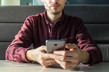Smiling male person with smartphone in cafe. Young man uses wireless device sitting at table in restaurant in bright daylight and waiting