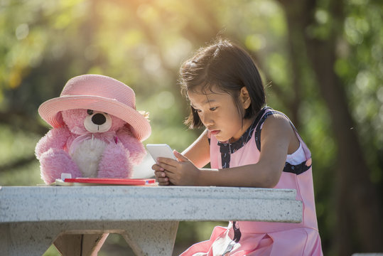 Little cute girl playing with teddy bear,she using smart phone.