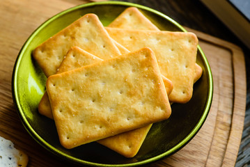 Salted crackers in green bowl on wooden tray. Part of book visible in one corner and small part of cream cheese spread on cracker in another corner.