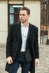 The solid man walks down the street. On him a black classical coat, a jacket and a white shirt. He looks in a distance, he has very serious look.