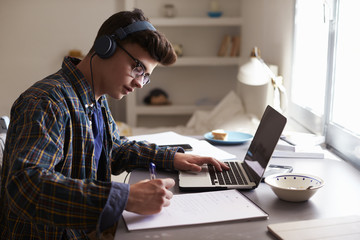 Teenage boy wearing headphones works at desk in his bedroom - Powered by Adobe