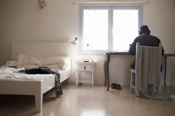 Teenage boy in headphones works at desk in bedroom, back view