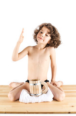 Cute curly-haired boy in shorts sitting in a "Hero of the position," bowed his head, raised his arm bent at the elbow. Before him is a wooden mug and towel. White background. © kravik93