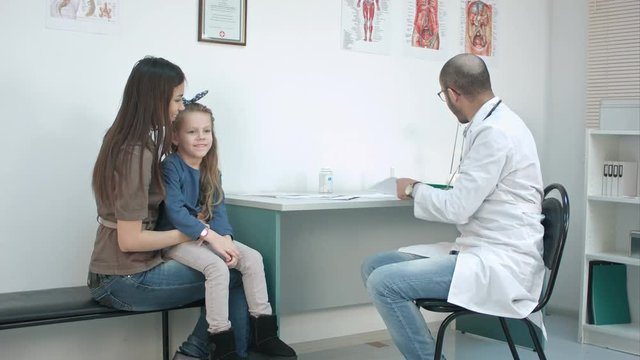 Little girl kissing her mom and smiling male doctor giving her candy for good behaviour