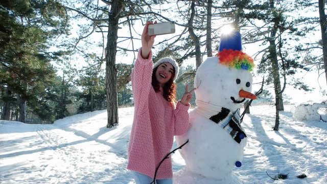 winter selfie, mobile phone in hand of a young woman making fun selfie photo in winter forest backlit, cute girl making photo with a snowman, snow sculpture, fun entertainment