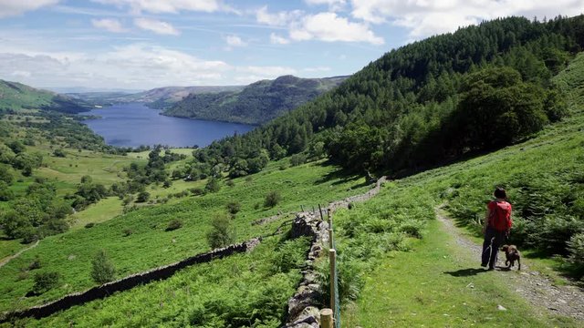 A hiker and their dog walking down a mountain path with views of Ullswater Lake on a sunny summers day from Glencoyne in the English Lake District, UK.