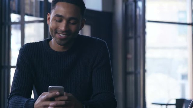 Smiling afro american guy sitting at cafe and typing message on mobile phone