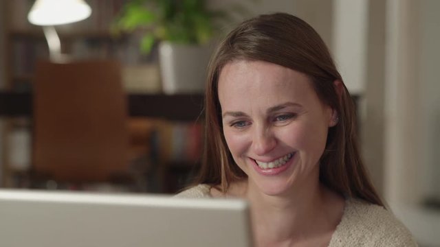 Young woman watches videos on internet in her laptop at home. She watches very concentrated and starts laughing with something very funny. Dolly shot.
