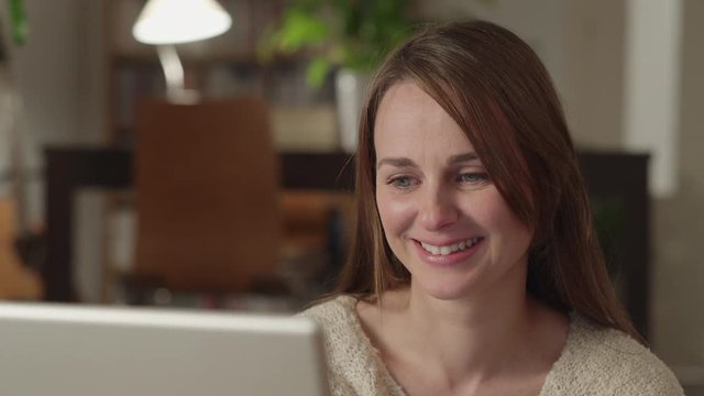 Young woman watches videos on internet in her laptop at home. She watches very concentrated and smiles. Dolly shot to the left.