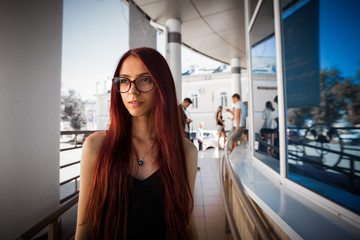 Urban city portrait of girl. Beautiful thinking girl with long red hair and glasses looking aside. Pretty girl in black dress on city, beautiful architecture background. Picture of pretty urban girl