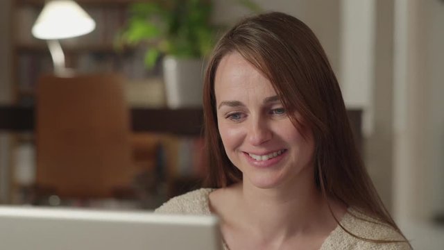 Young woman watches videos on internet in her laptop at home. She watches very concentrated and smiles. Dolly shot to the right.