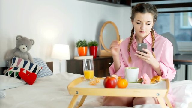 Happy girl in pyjamas eating orange in the bed and texting on smartphone
