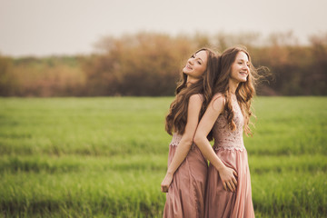 Beautiful happy young twins sisters in long evening dresses in green spring field in sunshine. Having fun together, positive emotions, bright colors. Copy space.