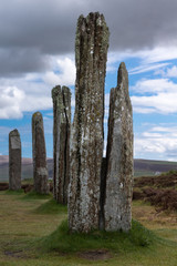Orkneys, Scotland - June 5, 2012: Ring of Brodgar Neolithic Stone Circle. Multiple gray menhirs with white and yellow mold spots stand erect on a grass field under blue and gray sky. Hills in back.