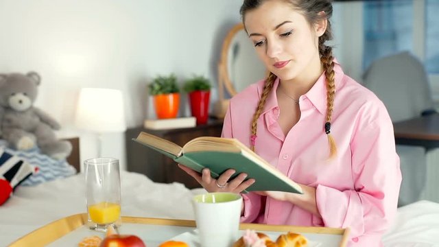 Girl finishes reading book and eats breakfast in bedroom, steadycam shot
