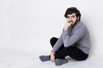 A portrait of handsome man having dark beard with moustache wearing casual clothes sitting on the floor crossed legs holding his hand on his chin looking into camera with mysterious look