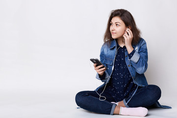 Naklejka premium People, lifestyle and technology. Fashionable young woman sitting crossed legs on white floor holding smartphone in her hands listening to the music typing messages looking aside being pleased