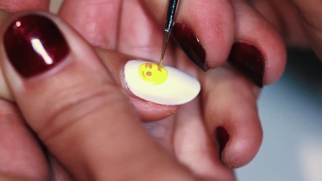 Manicure procedure woman hand draws yellow emoticon on white finger nail polish, close up