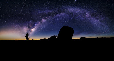 Photographer doing astro photography in a desert nightscape with milky way galaxy.  The background is stary celestial bodies in astronomy.  The heaven depicts science and the divine.