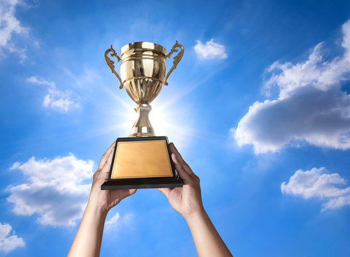 A Man Holding Up A Gold Trophy Cup With Bluse Sky And Sun Backgr