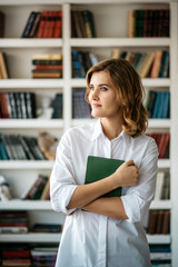 Beautiful young girl standing with book in hand in the library. Many shelves with books on a blurred background